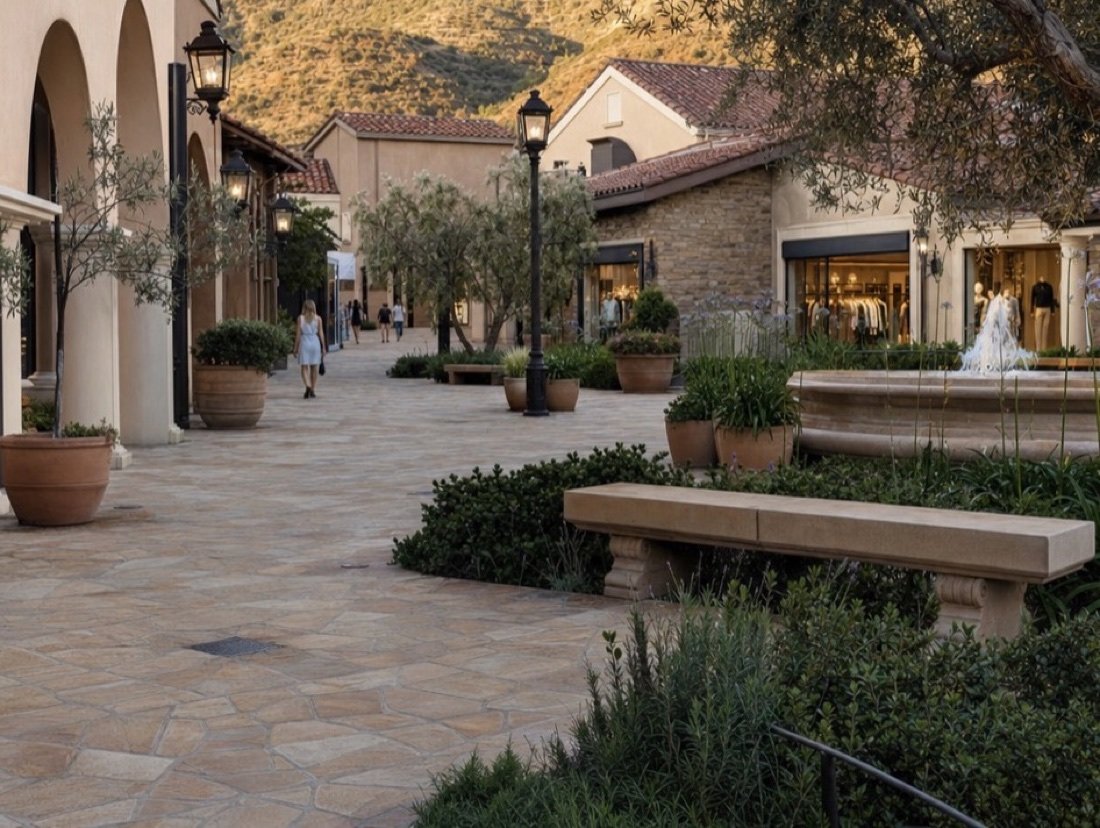 Shops under terracotta roofs and arched walkways around a stone-paved plaza with olive trees in planters and a shallow fountain
