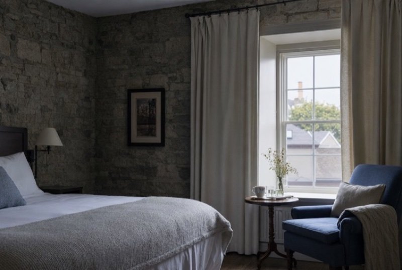 An inn bedroom built against rough gray limestone, the bed made and an armchair by the window, a calm corner of the hospitality industry